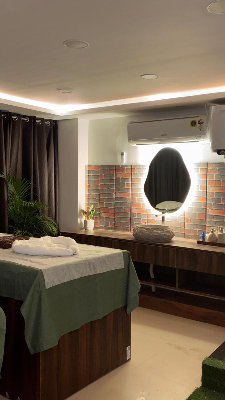 A treatment room: a bed in green linen, a backlit stone mirror on the red-brick wall, palms in the corner